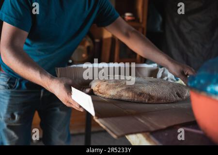 Front view of cropped anonymous local male chef in blue shirt standing at table with baked bread placed on carton in workshop Stock Photo