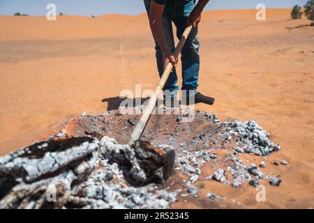 Cropped anonymous man using shovel while piling hot smoking charcoal in dig hole in sandy desert of Marrakesh Stock Photo