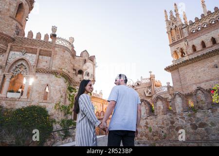 Side view of couple holding hands tourists standing on lawn of historic castle smiling and looking at each other against cloudless blue sky in daytime Stock Photo