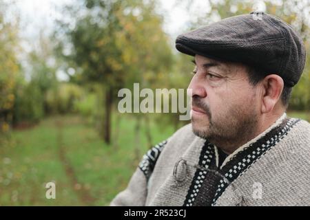 Thoughtful senior male in traditional Mapuche clothing and beret looking away while standing against blurred forest in Temuco Stock Photo