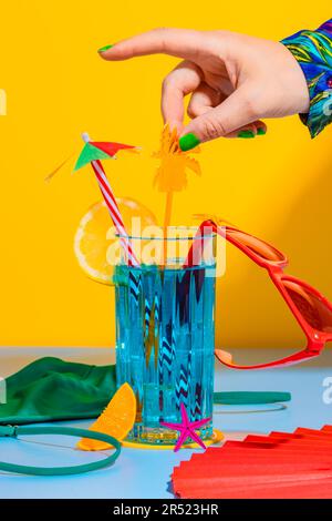 Cropped hand of anonymous person holding straw over glass of cocktail with sliced lemon, sunglasses next to inner wear on yellow and blue background Stock Photo