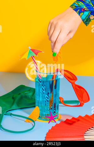 Cropped hand of anonymous person holding straw over glass of cocktail with sliced lemon, sunglasses next to inner wear on yellow and blue background Stock Photo