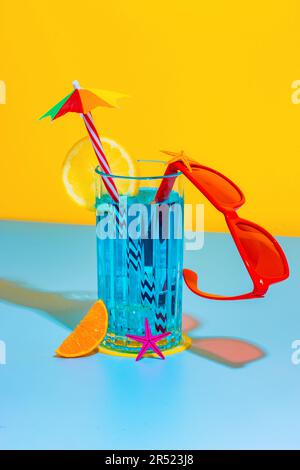 Composition of glass of blue liquid with slice of lemon and straw with stylish red eyeglasses placed on bright blue and yellow background in studio Stock Photo