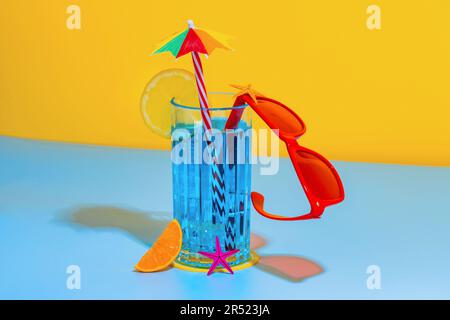 Composition of glass of blue liquid with slice of lemon and straw with stylish red eyeglasses placed on bright blue and yellow background in studio Stock Photo