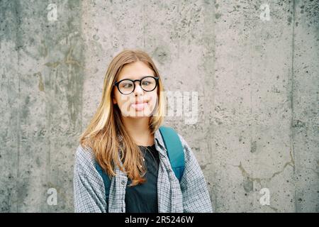 Outdoor portrait of silly young teenage kid girl pulling a tongue, crossing eyes over her nose, wearing glasses and backpack, posing on grey wall back Stock Photo