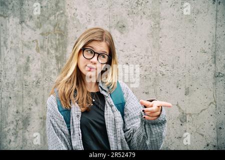 https://l450v.alamy.com/450v/2r52479/outdoor-portrait-of-young-teenage-kid-girl-wearing-glasses-and-backpack-posing-on-grey-wall-background-talking-and-pointing-finger-to-the-side-2r52479.jpg