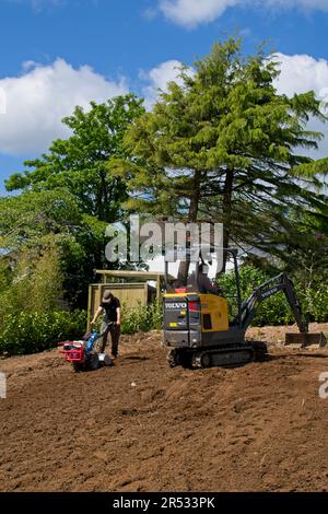 Gower Wales UK May 2023 Machinery preparing ground for replacement grass lawn. Mechanical digger and rotavator in use. Trees and bushes in background. Stock Photo