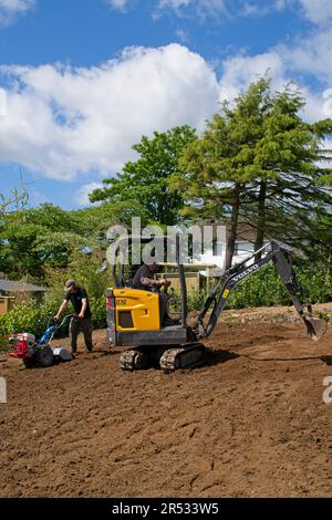 Gower Wales UK May 2023 Machinery preparing ground for replacement grass lawn. Mechanical digger and rotavator in use. Trees and bushes in background. Stock Photo