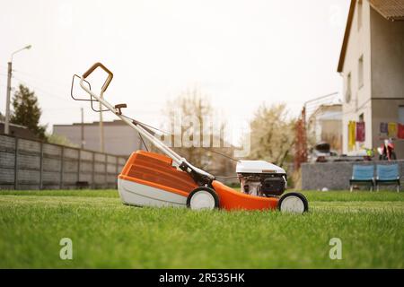 Modern orange-grey electric lawn mower on bright lush green lawn at residential backyard of house. Gardening work tools. Rotary lawn mower machine cut Stock Photo