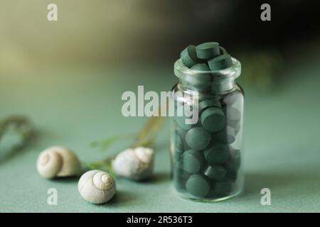 Spirulina pills in glass bottle, sea shells on green background. Antioxidant nutrition. Bio-energy, biofuel, energy research. Copy space Stock Photo
