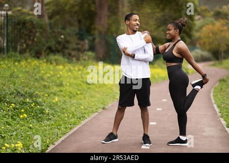 Positive young black couple with phone doing leg exercises, warm-up before running, enjoy sports Stock Photo