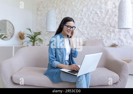 A young female student studies at home remotely on a laptop. He is sitting on the couch in a headset and jeans. Talks via video call, zoom meeting, listens to online lectures and lessons. Stock Photo