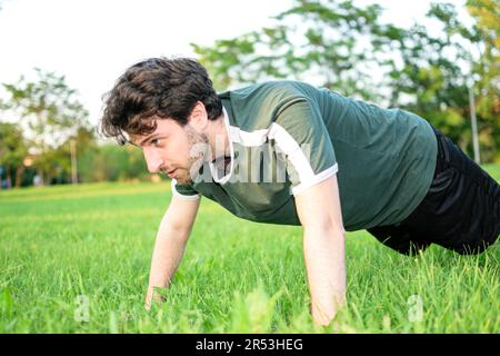 Young man with green t-shirt doing push-ups concentrated in park with grass and sunny day Stock Photo