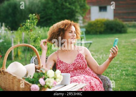 Outdoor portrait of beautiful mature woman resting in summer garden, sitting in cosy chair, having video call with her family or friend, basket with f Stock Photo