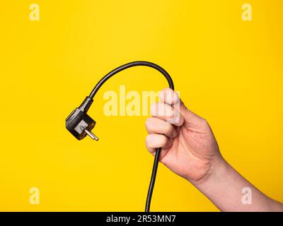 A hand holds a plug to which a black cable is attached. No face, yellow background. Stock Photo