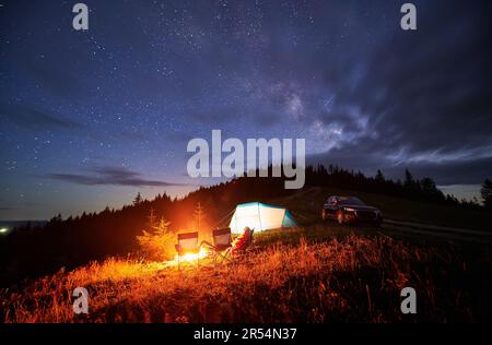 Cozy camping in mountains under evening cloudy sky. Starry night above bonfire, tourist tent and off-road car. Summer evening on hills. Stock Photo