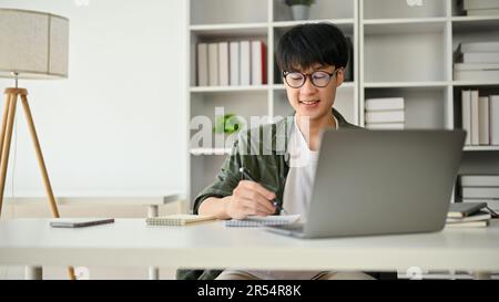 A smart and handsome young Asian male office worker is taking notes on a notepad and focusing on his work at his desk in the office. Stock Photo