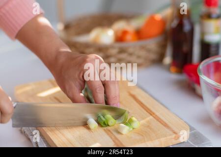 Female hand using a knife to cut Negi (Japanese Long Onion) or Japanese Scallion or Green Onion on wooden cutting board. Woman preparing food in the k Stock Photo