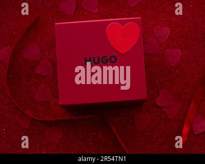 Hugo by Hugo Boss. Red gift box is on red heart paper background, top view. Greeting card, present. Valentines day holiday concept. Flat lay with a lo Stock Photo