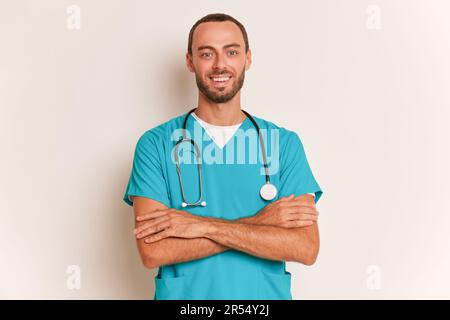 Bearded young medical worker wearing turquois medical uniform with stethoscope on his neck poses inside, holds his hands across his chest Stock Photo