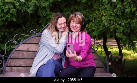 Grandmother in headphones with a phone listens to music. together with a friend on a park bench and listening to music. Stock Photo