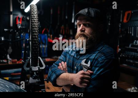 Side view of bearded cycling repairman in cap holding wrench in hand standing by bicycle in repair workshop with dark interior, serious looking at Stock Photo