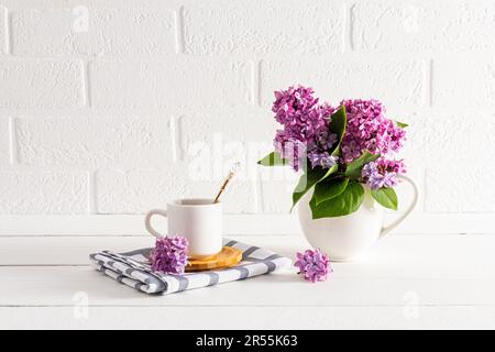 Beautiful spring still life with a cup of coffee and a jug with a bouquet of lilacs on a white wooden table. front view Stock Photo