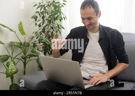Deaf or hard hearing happy smiling young caucasian man uses sign language while video call using laptop while sitting on the couch at home. Stock Photo