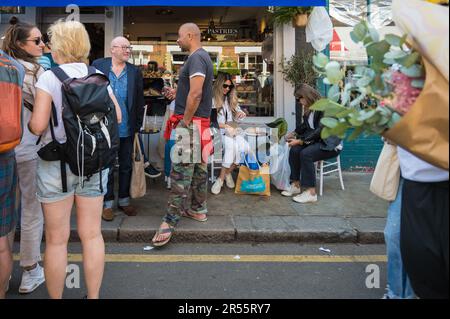 People queueing and in conversation outside cafe on market day at Columbia Road flower market. Two young women sit at pavement table. London, England Stock Photo