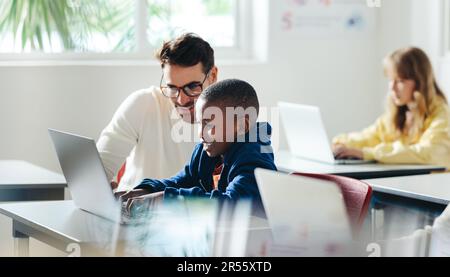 Male teacher helps a young boy with computer-based learning in a classroom setting. Child tutor providing a lesson in an elementary school, with a foc Stock Photo