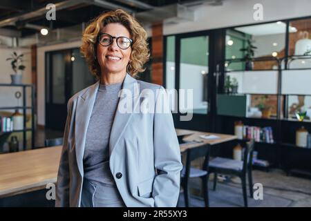 Portrait of a professional woman in a suit standing in a modern office. Mature business woman looking at the camera in a workplace meeting area. Stock Photo