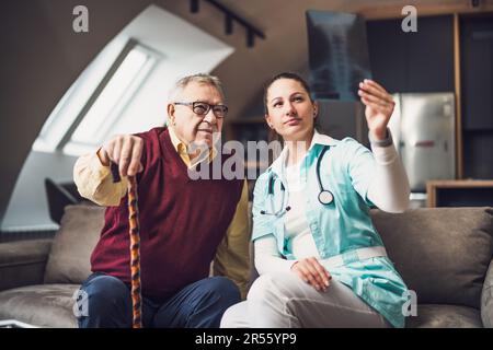 Home doctor is examining x-ray image with old man at his home. Professional health support for elderly people. Stock Photo