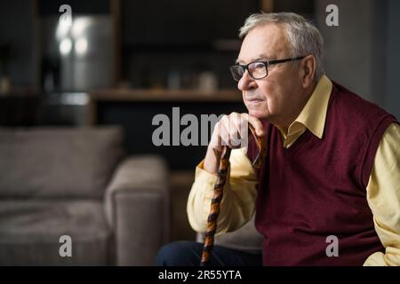 Pensive and worried senior man is sitting on bed in his home. Portrait of senior man with walking cane. Stock Photo