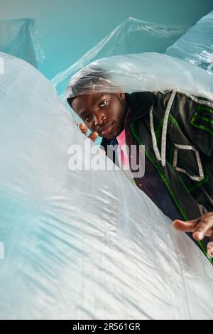 Portrait of confident young afroamerican male model in outwear jacket with led stripes looking at camera near glossy cellophane on turquoise backgroun Stock Photo