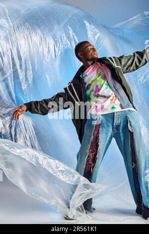 Trendy young afroamerican model posing in outwear jacket with led stripes and ripped jeans while standing on glossy cellophane on blue background, urb Stock Photo