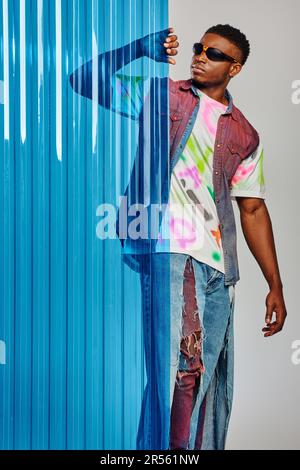 Good looking young afroamerican model in sunglasses, denim vest and t-shirt touching blue polycarbonate sheet and standing on grey background, fashion Stock Photo