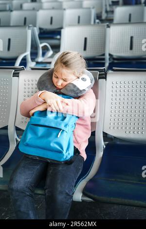 Kid, teen tired girl sleeping, waiting in airport passenger terminal departure hall with backpack. Sitting on chairs in airplane travel pillow. Flight Stock Photo