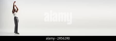 Full length of young african american man in sleeveless t-shirt and pants raising hands on grey background, contemporary shoot featuring stylish attir Stock Photo