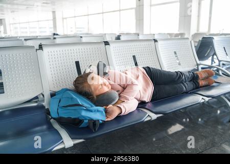 Kid, teen tired girl sleeping, waiting in airport passenger terminal departure hall with backpack. Sitting on chairs in airplane travel pillow. Flight Stock Photo
