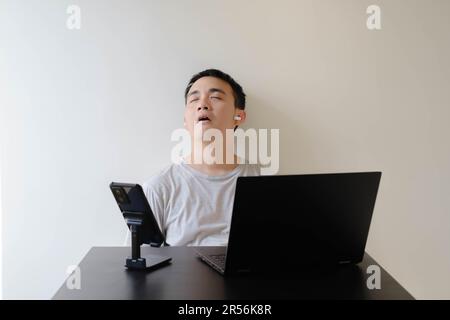 A young Asian man wearing a grey t-shirt and a pair of wireless earphones is falling asleep while working from home Stock Photo