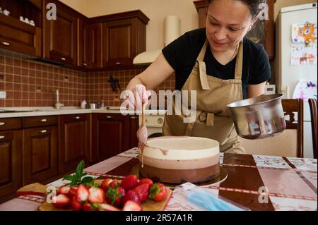 Pregnant female chef confectioner decorating with ruby pink glaze the birthday cake with three chocolate mousse layers Stock Photo