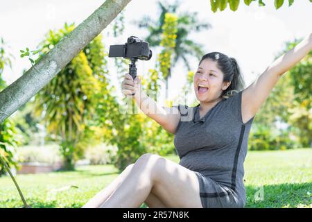 young latin influencer woman sitting on the grass of the park recording a video with great happiness while raising her hands and smiling. Stock Photo