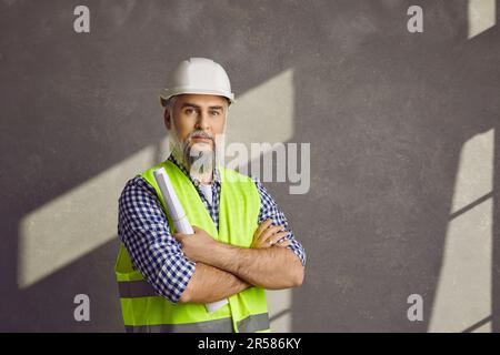 Portrait of mature bearded foreman in a helmet and construction vest on grey wall background. Stock Photo