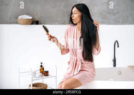 Puzzled surprised caucasian woman in a bathrobe, with thick long black hair, sits in the bathroom, holds a comb in her hand, looking at it in amazement after brushing her hair Stock Photo