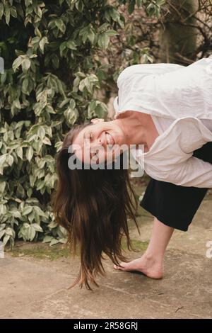 Happy smiley female yoga teacher in bound extended side angle in leafy terrace of landscaped garden. Looking at camera. Showing her tongue. Wearing bl Stock Photo