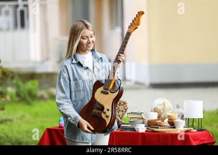 Woman holding guitar near table with different items on garage sale in yard Stock Photo