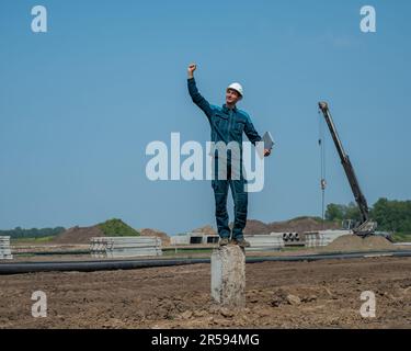 Builder in a protective helmet holds a laptop and stands on a pile in the middle of a construction site.  Stock Photo