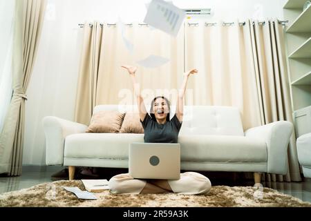 Asian woman throws papers in the living room as she finishes her work with her laptop on her lap. Stock Photo