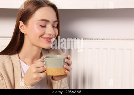 Woman holding cup with hot drink near heating radiator indoors Stock Photo