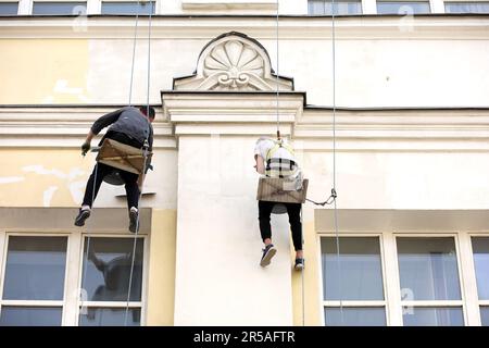 Workers paints the building wall. Painters hanging on a cable with paint buckets, steeplejack repairing old house facade, renovating works Stock Photo